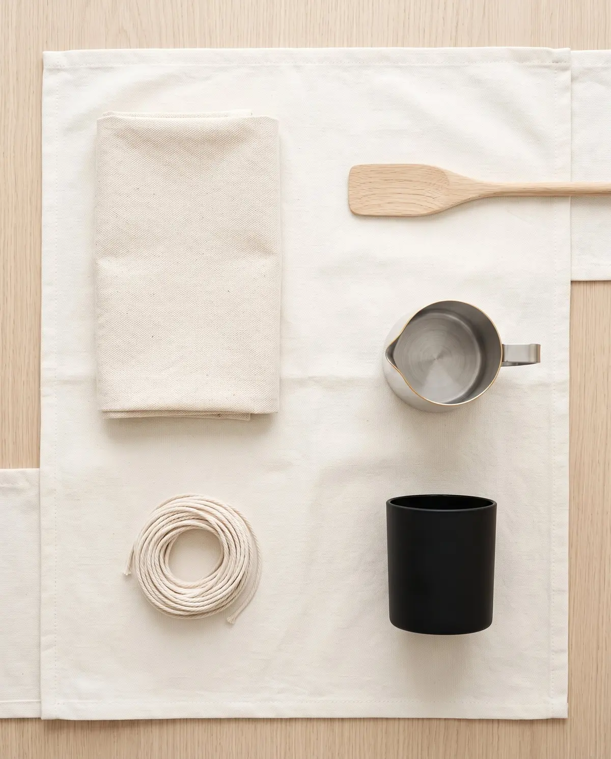 A workshop flat-lay from above: a folded cotton cloth, a wooden spatula, a steel pouring jug, a coil of cotton wick and an empty matte black vessel on canvas.